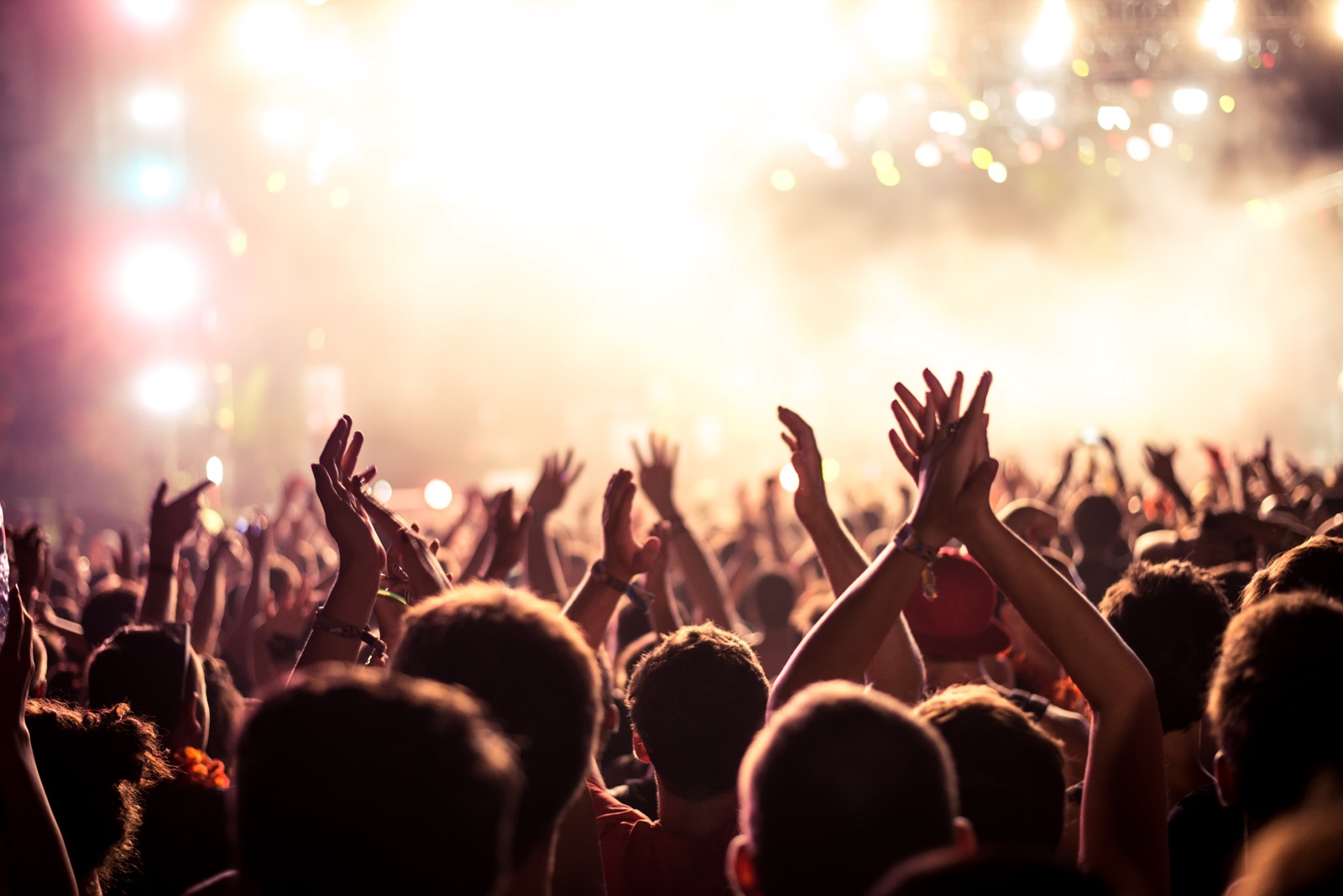 A crowd with arms raised and hands clapping, silhouetted against bright stage lights.